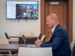 Adult student doing schoolwork on laptop in Cole Hall, Atrium with digital screen behind him promoting University events.