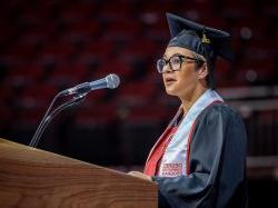 Female degree completion student speaking at Commencement in black regalia with white "Degree Completion Program" graduation stole.